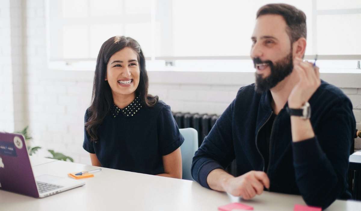 two coworkers sitting at a desk with a laptop interacting